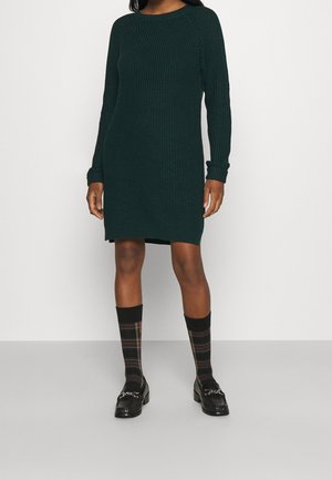 Femme portant une robe en tricot vert foncé à manches longues, des chaussettes hautes à carreaux noires et des mocassins noirs sur un fond clair uni.