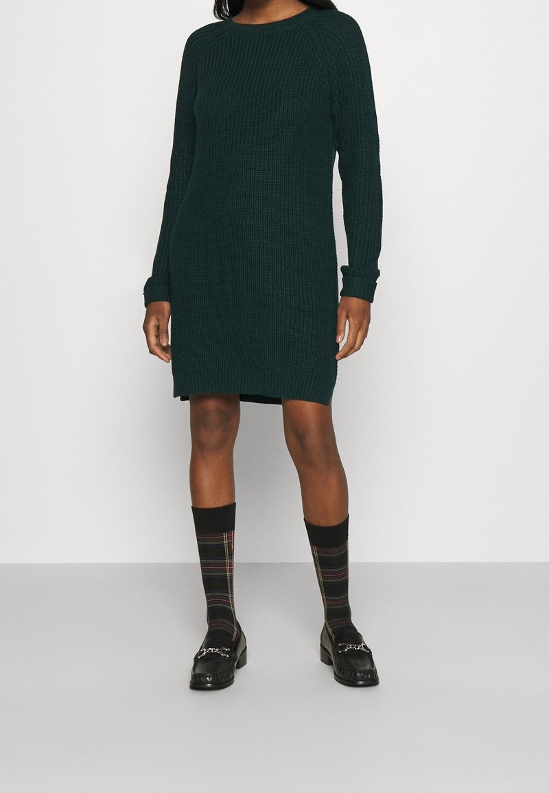 Femme portant une robe en tricot vert foncé à manches longues, des chaussettes hautes à carreaux noires et des mocassins noirs sur un fond clair uni.
