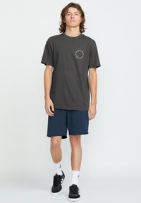 T-shirt en coton gris foncé à manches courtes, présentant un logo blanc circulaire sur la poitrine. Associé à un short bleu marine et des baskets noires.