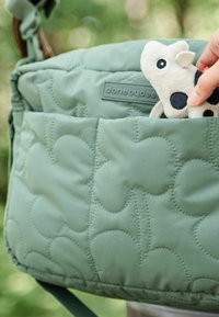 Képtextúrájú zöld táska, sima felülettel, logós címkével, és egy plüss tehén játék részben látható az elején lévő zsebében.