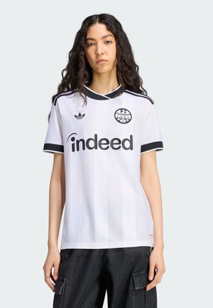 Kobieta z długimi kręconymi włosami ubraną w białą koszulkę piłkarską Eintracht Frankfurt z czarnymi lamówkami i czarne spodnie typu cargo.