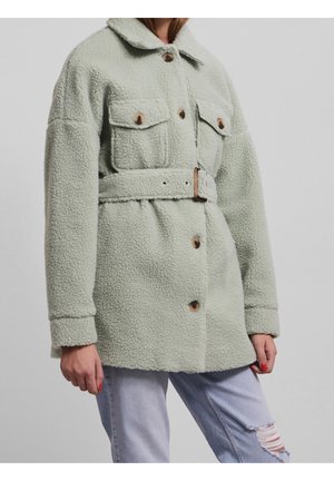 Vrouw draagt een lichtgroene teddyjas met riem, grote knopen en borstzakken, gecombineerd met lichtblauwe gescheurde jeans.