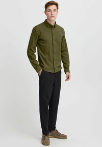 Olive green button-up shirt with a collar, long sleeves, and a smooth texture, paired with black trousers and brown footwear.