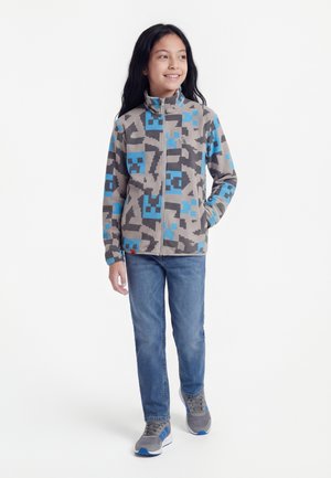 Jeune fille souriante, portant une veste zippée grise et bleue à motifs, un jean bleu et des baskets grises, debout devant un fond blanc.