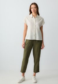 Donna in piedi che indossa una camicia a maniche corte beige chiaro con bottoni, pantaloni verdi oliva cropped e sneakers bianche su uno sfondo neutro.