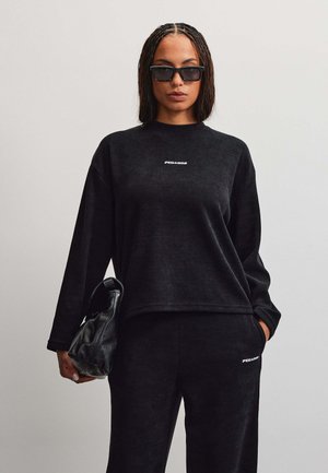 Donna che indossa top nero a maniche lunghe e pantaloni con scritta bianca, occhiali da sole neri, tiene una borsa in pelle nera, su sfondo grigio uniforme.