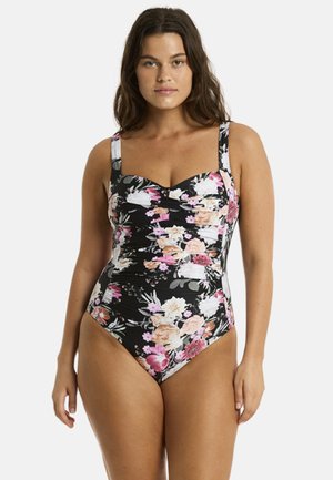 Frau trägt einen schwarzen Badeanzug mit pinkem, weißem und pfirsichfarbenem Blumenmuster und steht vor einem schlichten hellen Hintergrund.