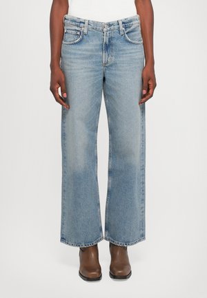 Jeans de pierna ancha en un azul claro de material de mezclilla desgastado. Con cinco bolsillos y una clásica tapa de botón y cremallera. Combinados con botas marrones.