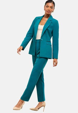 Kvinde poserer i en skræddersyet teal-dragt med matchende blazer og bukser, cremefarvet top og beige højhælede sko, foran en hvid baggrund.