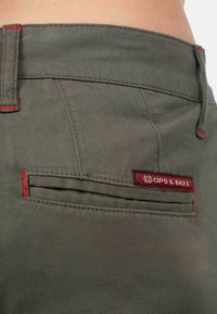 Olijfgroene stoffen broek met rode stiksels, voorzien van een zijzak en een bordeauxrood label met de merknaam "Cipo & Baxx."