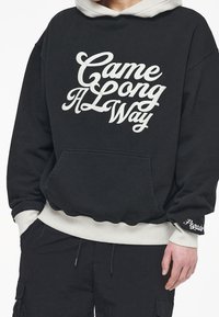 Persona che indossa una felpa nera con polsini e orlo bianchi, con la scritta "Came a Long Way" sul davanti e "Pegador" ricamato sul polsino sinistro.