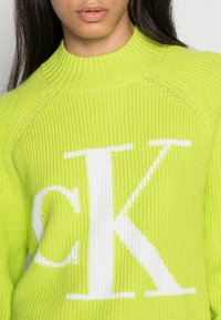 Helles limettengrünes Strickpullover mit hohem Kragen. Verfügt über ein großes weißes "CK"-Logo. Strukturierte Rippung ist im gesamten Stoff sichtbar.