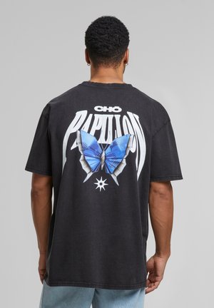 Svart bomull t-skjorte med et stort blått sommerfugl-grafikk, omgitt av hvit tekst "CHO PAPILLON" på baksiden. Løs passform med korte ermer.
