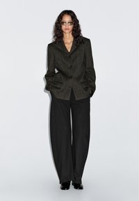 Kobieta stoi przy gładkiej ścianie, ubrana w oversize'ową, ciemnoszaro wzorzystą marynarkę, szerokie czarne spodnie, czarne buty i przyciemniane okulary przeciwsłoneczne.