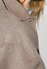 Brun uldfrakke med spidse kraver, sorte knapper og en struktureret overflade, taget fra en sidevinkel.