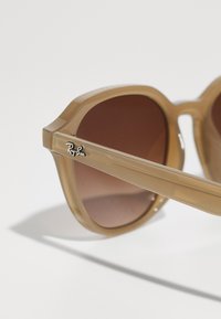 Ochelari de soare cu nuanță maro, având o ramă translucidă de culoare bej, cu o formă octagonală și un mic logo Ray-Ban argintiu pe tâmplă.
