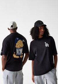 T-shirt noir avec un graphique d'un seau de popcorn sur le dos et un logo LA blanc à l'avant. Tissu en coton doux, coupe oversize.