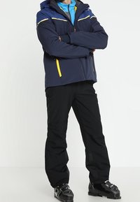 Marineblauwe skijas met gele accenten, twee borstzakken en een capuchon, gecombineerd met zwarte broek en skischoenen. Model staat met de armen gekruist.