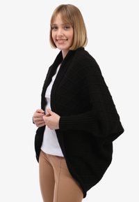 Scialle nero lavorato a maglia con un motivo a diamante testurizzato, con apertura frontale e maniche flessibili. Indossato sopra una maglietta bianca e pantaloni beige.