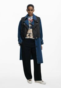 Trench coat in denim con sovrapposizione di un gilet in pelle nera, con colletto a risvolto, zip e tasche. Indossato sopra una camicia a fantasia e pantaloni neri.