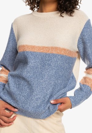 Frau trägt einen weichen Strickpullover mit cremefarbenen, orangefarbenen und blauen Farbfeldern, Hände ruhen nahe der Taille, teilweise sichtbares lockiges Haar.