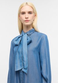 Camicia di jeans azzurro chiaro con colletto e un dettaglio a papillon, presenta maniche lunghe e una trama morbida e liscia.