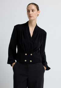 Sort indpakningsbluse med lange ærmer og plisseret design. Har guldtonede knapdetaljer i taljen. Blødt, glat stof.