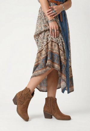 Kvinde iført mønstret asymmetrisk nederdel, denimskjorte, brune ruskindsstøvler med stablet hæl og flere sølvringe og armbånd på hænderne.