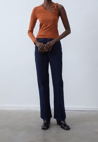 Langer, rostfarbener Langarm-Top mit einer gedrehten Schulterdetails, kombiniert mit dunkelblauen, ausgestellten Jeans und schwarzen Stiefeletten. Glatte Textur.