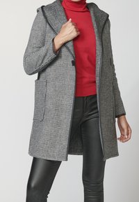 Cappotto a scacchi grigio e nero con un ampio cappuccio, tasche frontali e finiture nere a contrasto, indossato sopra un dolcevita rosso e pantaloni neri.