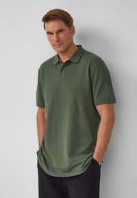Olijfgroene polo met kraag, voorzien van drie knopen en korte mouwen; de stof heeft een gladde textuur en een relaxte pasvorm.