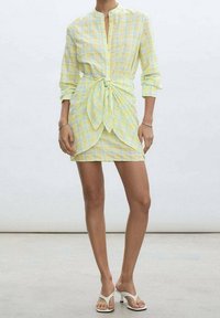 Světle žluté a zelené kostkované šaty s uzlem v pase, dlouhými rukávy, knoflíkovým zapínáním vpředu a hladkým povrchem. Doplnit s bílými sandály.