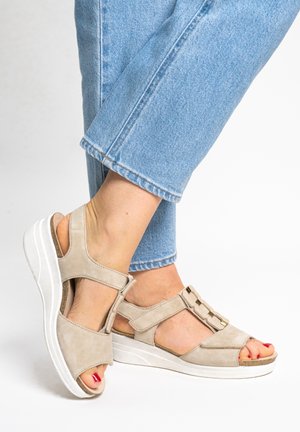 Feet wearing beige suede wedge sandals with ankle straps and gold chain detail, paired with light blue cropped jeans.