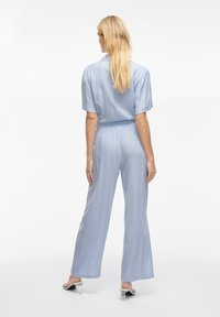 Gaiši zils kombinezons ar īsām pūkainām piedurknēm, elastīgu jostasvietu un platiem zobiem. Izgatavots no mīkstas, plūstošas auduma ar gludu tekstūru.