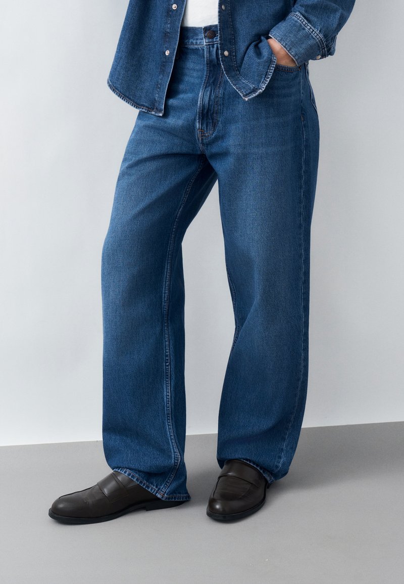 Άτομο που φοράει φαρδιά μπλε τζιν, σκουρό καφέ παπούτσια χωρίς κορδόνια και ταιριαστό μπλε τζιν μπουφάν, με το ένα χέρι στην τσέπη.