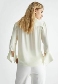 Blusa color crema con maniche tre quarti, dotata di polsini a spacco, texture liscia e vestibilità morbida. Design minimalista senza motivi.