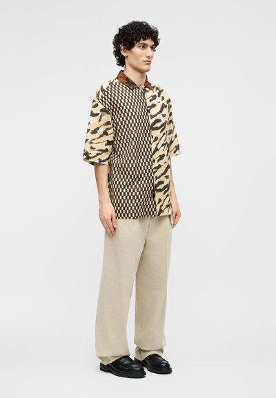 Νέος άνδρας όρθιος, φορώντας πουκάμισο με κοντά μανίκια και πατς εργασίας με σχέδια σκακιέρας και ζώων, μπεζ παντελόνι με φαρδιά πόδια και μαύρα παπούτσια.