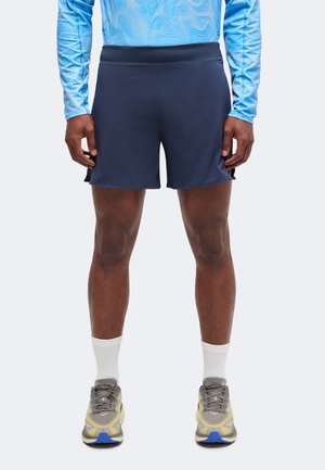 Personne portant un short de sport bleu marine, un t-shirt à manches longues bleu clair, des chaussettes blanches et des chaussures de course grises, debout.