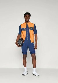 Kolesarska oprema, ki vključuje oranžni brezrokavnik z modrim napisom in tesno prilegajoč jumpsuit v mornarsko modri barvi. Vsebuje črno čelado, bele čevlje in eleganten dizajn.