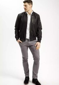 Czarna kurtka bomberka z kieszenią na zamek, noszona na białej koszuli, połączona z szarymi spodniami i czarnymi butami, na jasnym tle.