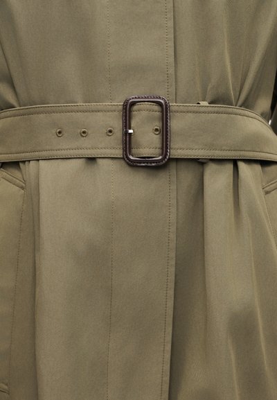 Κοντινό πλάνο καμπαρντίνας από ελιάς με ραμμένη ζώνη και ορθογώνια σκούρα καφέ αγκράφα στερεωμένη στη μέση.