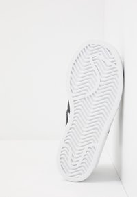 Baskets noires montrant une semelle en caoutchouc texturée avec un motif en chevron, des bords blancs et un design épuré, vues par le dessous.