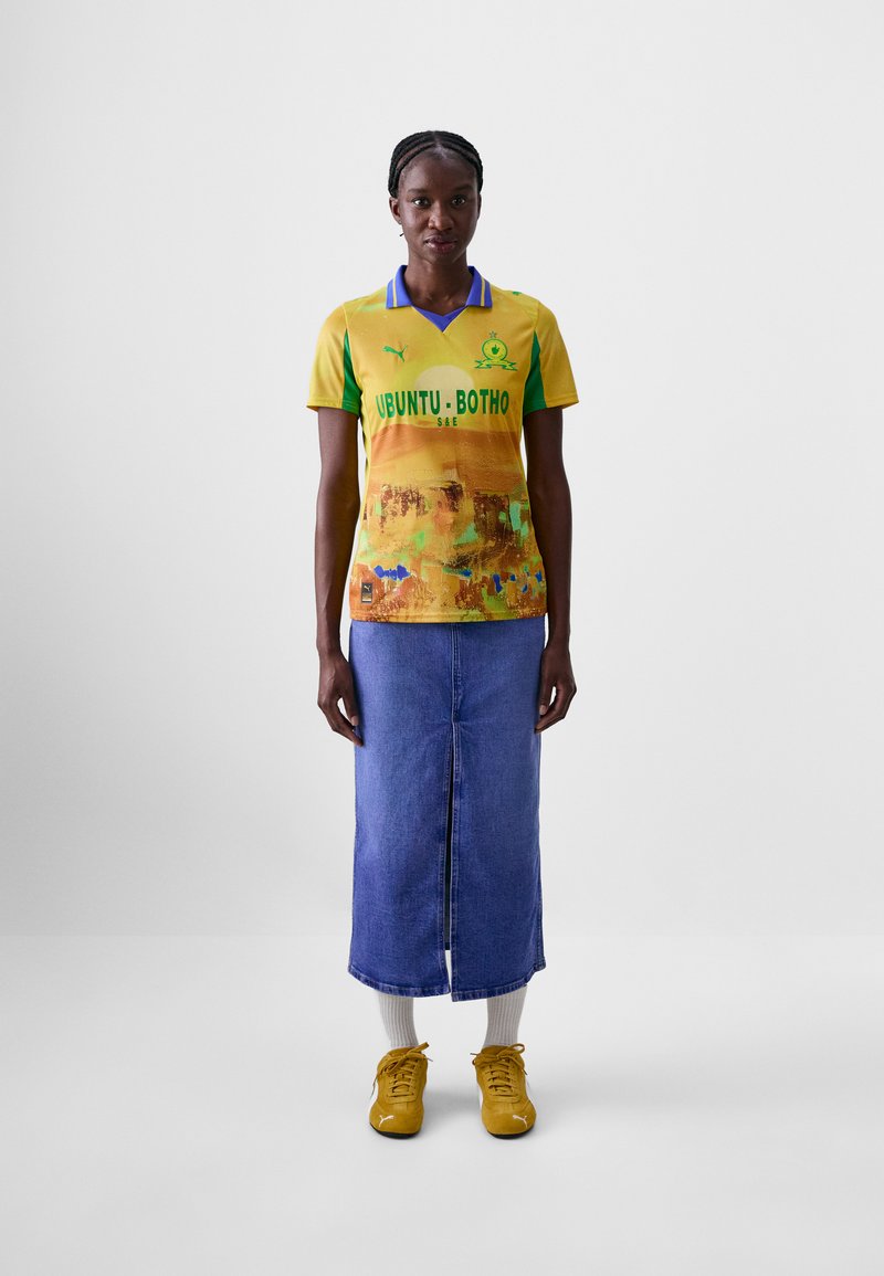 Maglietta gialla e verde con design astratto, con il testo "UBUNTU-BOTHO". Indossata con una lunga gonna di jeans e sneakers gialle.