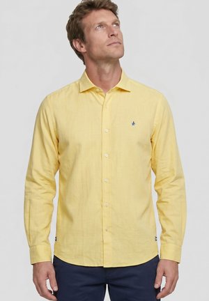 Hombre con camisa amarilla de manga larga abotonada y pantalones oscuros, mirando hacia arriba con un fondo claro y sencillo.