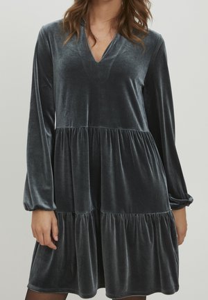 Femme portant une robe longue en velours gris foncé à manches longues, col en V et jupe à volants froncée, debout devant un fond clair uni.