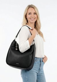 Blonde vrouw in witte blouse en blauwe spijkerbroek, glimlachend, met een grote zwarte leren schoudertas met metalen bedels.