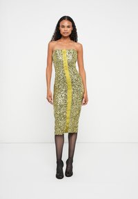 Nő, aki pánt nélküli, arany flitteres midi ruhát visel, középen fényes csíkkal, fekete áttetsző harisnyával és fekete nyitott orrú magassarkú cipővel áll egyenesen.