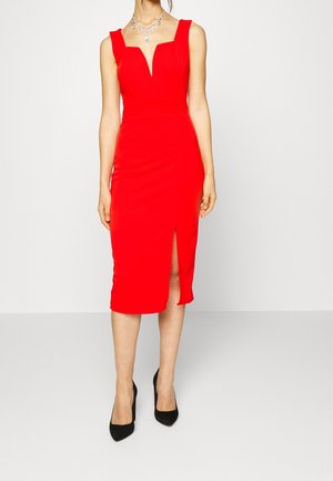 Vrouw draagt een aansluitende rode mouwloze midijurk met een voorste split, gecombineerd met zwarte pumps met spitse neus en een zilveren ketting.