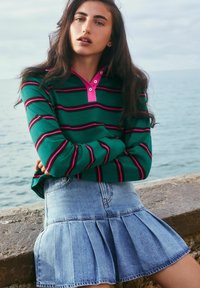 Green striped pullover with pink accents and buttons, paired with a light blue pleated denim skirt. Background features a water view.