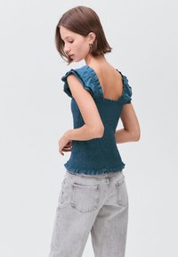 Mlada žena s kratkimi rjavimi lasmi, oblečena v turkizno ruffled brezrokavno bluzo in svetlo sivo jeans hlače, stoji ob enobarvni svetli podlagi.